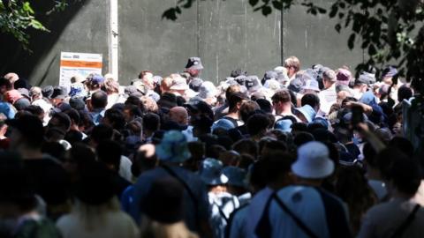 A large crowd of unidentifiable people stands around outside a barricaded area. Many are wearing bucket hats. 