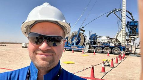 A man with protective goggles, a blue top, and safety helmet stands in front of machinery on an oil field