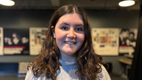Headshot of 16 year-old girl with long brown hair smiling at the camera. She is wearing a grey woolen turtle neck. The background of the room she is in is blurred but posters on culture are visable.