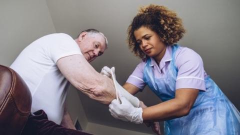 A carer puts a bandage on an old man's arm while he sits on a sofa