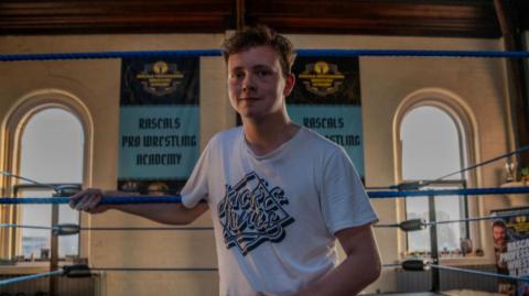 A young man in a white t-shirt standing in a boxing ring smiling.