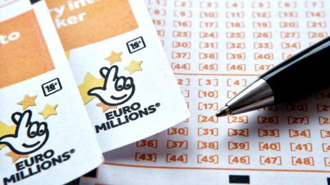 Two EuroMillions tickets side-by-side, with the National Lottery logo seen at the top of both, beside a ballpoint pen