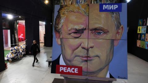 Giant painting showing composite face of Trump and Putin divided into quarters with the word peace in Russian and English at the top right and bottom left corners. It stands in a gallery in St Petersburg and the artist, Alexei Sergienko, is seen walking to the left of the picture.