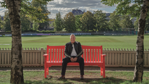 Lee sitting on a red bench framed by two tree trunks in the foreground and with a cricket pitch behind him surrounded by mature trees. He is smiling and wearing a black track suit