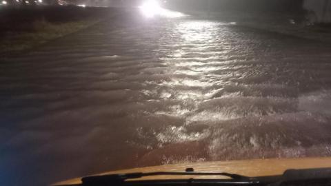 View of a flooded road at night taken from inside a vehicle. The white bonnet and windscreen wipers are visible at the bottom of the screen. Deep water lit up by street lights stretches out into the distance.