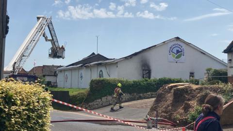A white rendered social club during a fire, with its roof completely gutted and smoke damage to its exterior. There is no physical smoke and there is an aerial appliance with a couple of fire fighters hovering above the building. White and orange tape has been strung across the road and a fire fighter can be seen walking towards the building. The sky above is blue with a few small clouds.