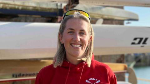 Emma is wearing a red 'Jersey Rowing Club' hoodie standing in front of stacked kayaks by a waterfront, with calm water, stone wall, and distant buildings under a sunny sky.