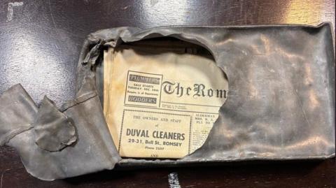 A large lead box is pictured with part of its front prised open. Inside is a newspaper, slightly faded, which is from 1969. 