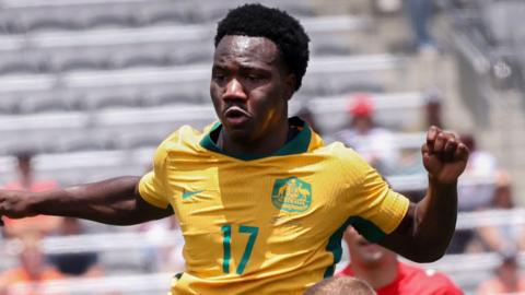 Nestory Irankunda, wearing Australia's yellow home shirt with green trim, challenges for an aerial ball in a World Cup warm-up game against Switzerland