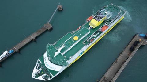 A birds-eye view of a DFDS ferry docking in a Jersey port. The ferry has green flooring, with white and yellow sides and there is smoke coming from its chimney.