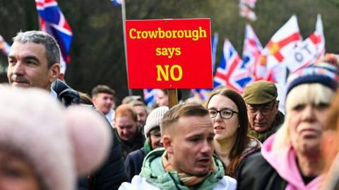 People take part in a demonstration in Crowborough. A red sign reading "Crowborough says NO" is held above the crowd, with Union flags visible behind it.