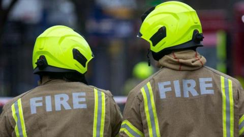 The backs of two firefighters with bright yellow helmets and wearing brown firefighting outfits that say "fire" on the back.