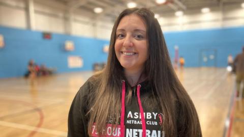 A woman with long brown hair smiling at the camera. She is wearing a pink lanyard and top which has "Ashby Netball Club on it. Behind her is a sports court. 