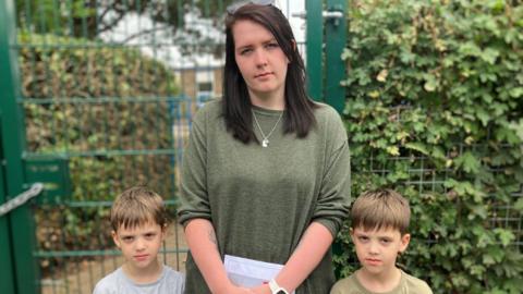 Danielle Hall and her young twin sons wear serious expressions as they stand by a fence. She has long, straight, dark brown hair and wears a green, long-sleeved T-shirt. The boys have brown hair and T-shirts.