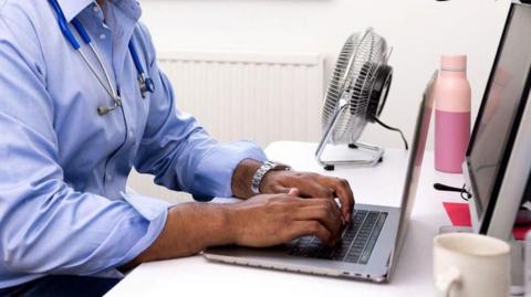 A man wearing a blue shirt sitting at a desk using a laptop. He is a GP and there is a fan at his desk.