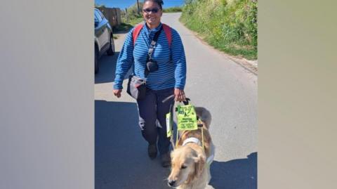 Lena Welch is walking with her guide dog. Lena is wearing a blue stripey long sleeve top. The Labrador Retriever mix is golden in colour and has a high visibility harness on. They are walking down a road with some greenery on either side and a car parked on the left.