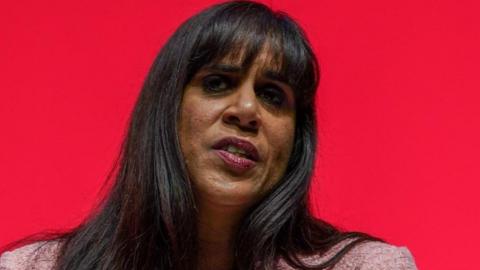 A woman with long dark hair is wearing a pink jacket. She is in front of a red background.