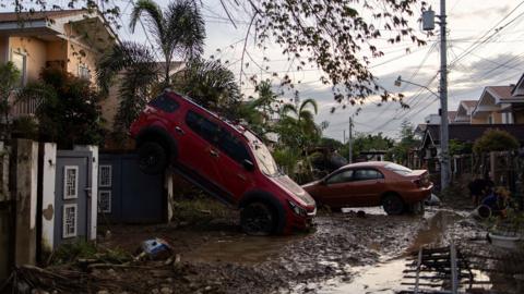 A car's back wheels sit on a toppled over house.