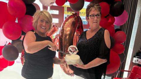 A blonde woman in a black shirt standing next to a woman with brown hair who is wearing a black shirt. There are balloons behind them and they are cutting a cake which says "Lee's 40th anniversary" on top.