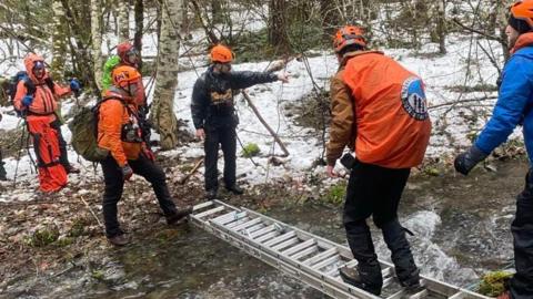 Volunteers seen crossing a creek on a ladder in a snowy forest