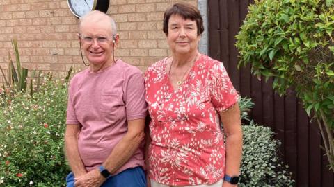 Pete wearing a pink T-shirt and blue shorts standing next to Sue wearing a pink and white floral shirt and white trousers - both standing in their back garden 