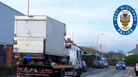 A street with industrial units on the left side and a residential house surround by a hedge on the left. In the foreground there is a white truck with a trailer that has a white van on the back. In the background there are four cars parked on the left and one blue car driving towards the camera on the right. A woman is just visible on the right of the frame