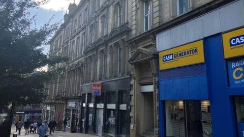 A four-storey Victorian-era bank building sits between a Greggs store and a Cash Generator pawnbrokers.