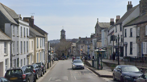 Lower Market Street in Penryn on a bright day. The road has road humps near a bus stop and there are cars parked on both sides. At the end of the street is a clock tower with a rounded dome and cross. The buildings lining the pavements on both sides are three and four storeys and some have shop windows on the ground floor.