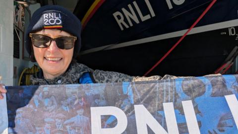 Anjie Rook holding up a blue RNLI flag in front of an RNLI vessel. She is wearing sunglasses and a navy blue hat which reads RNLI 200.