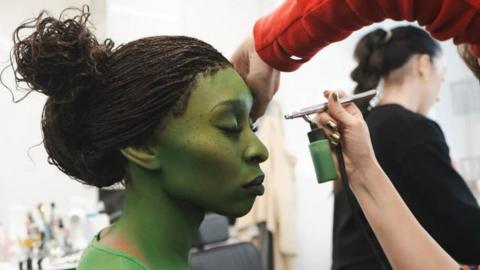 Cynthia Erivo sat in a make-up chair in a make-up trailer on the set of Wicked. She has her eyes closed and is being painted green by someone who is using an air brush machine. 