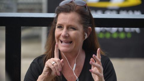 A woman signing, she has a black t-shirt and has earphones in.