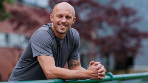 Jack Higgins leans on a metal fence. He is wearing a grey tee shirt. There is a brick building behind him in the background.