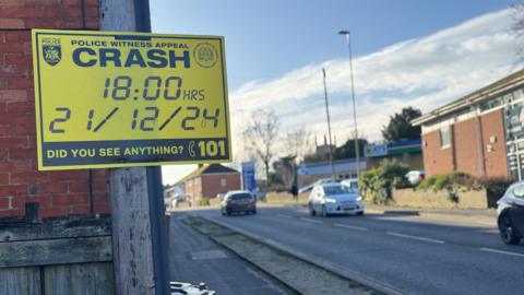 A yellow and black police sign on a post on the pavement. A road with cars and a petrol station can be seen in the background.