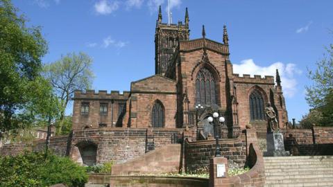 St Peter's Collegiate Church in Wolverhampton is a large medieval church built from red bricks, photographed on a sunny day against a blue sky. Stone steps lead up to the entrance. There are ornate lamp posts and a statue on the right-hand side. The church's tower rises above the building, surrounded by green trees.