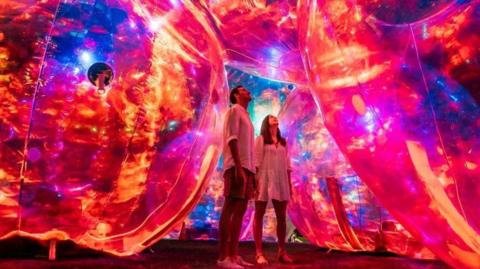 Giant glowing bubbles lit up in a red, orange and blue light. Two people are stood in the middle looking up.