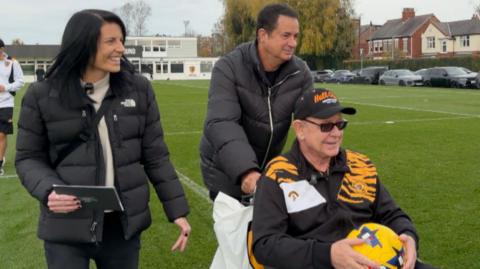 Hull City owner Acun Ilicali, a man in long black coat, pushes Owen Griffiths, an elderly man wearing a Hull City tracksuit and holding a yellow football, in his wheelchair. A woman with dark hair and wearing a black jacket smiles as she walks beside them.