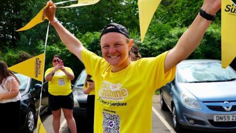Laura Bird standing with her arms aloft holding yellow bunting and smiling broadly. She is wearing a yellow T-shirt saying Rays of Sunshine. Behind her are parked cars and a woman in a yellow T-shirt taking a pic of her. 