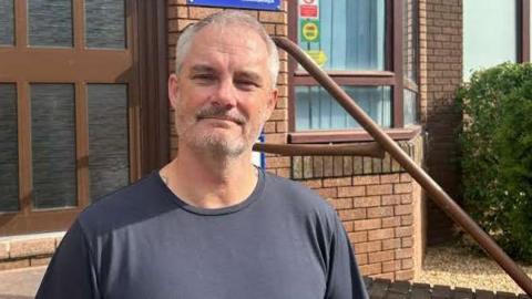 Alan Dobson standing on the steps outside the We Are With You offices. He has cropped grey hair and a short stubble beard and is wearing a black t-shirt