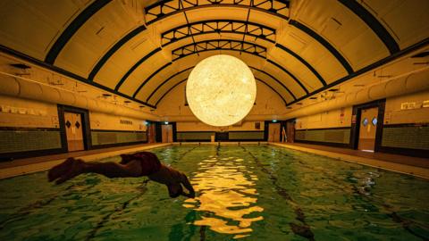 A silhouette of a swimmer diving into an indoor pool. There is a large sculpture modelled off of the sun suspended above the pool. It is reflecting in the water.