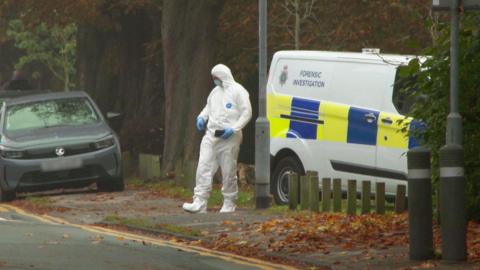 A forensic investigation officer wears a white suit that covers their head and feet, with blue gloves. They are standing next to a Staffordshire Police forensic invest van with blue and yellow livery