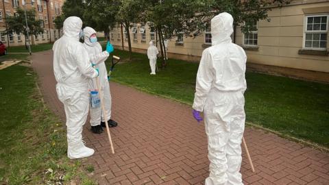 Three police officers wearing full-body white protective suits stand on a brick pathway bordered by grass. They are near blocks of flats with rows of windows. There are trees beside the path, which runs between the buildings. Two of the officers have poles they have been using to search the area. The third officer has a spade.
