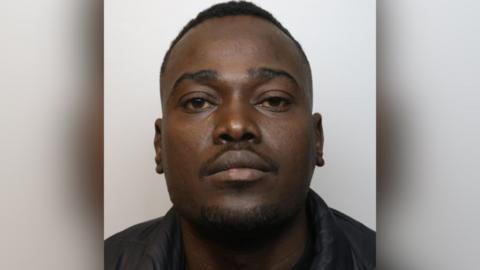 A mugshot of Abdulmawal Ibrahim Adam. He has short dark hair and stubble on his chin and upper lip. He is pictured looking directly at the camera with a blank expression.