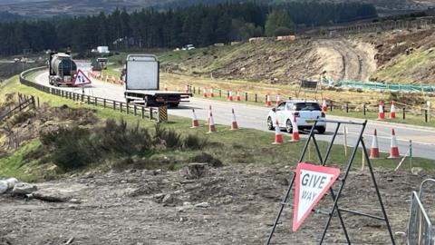 A9 with a queue of moving lorries and cars. There are traffic cones along the side of the road and new tracks can be seen for construction vehicles. There is a give way triangular sign next to the road.  