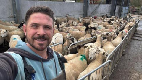 Joshua takes a selfie, while standing in front of a pen filled with sheep in his farmyard