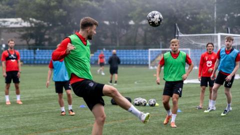 A man kicks a football in the air on a green pitch. Other players in green and blue bibs look on, with two goals in the distance.