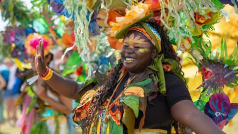 A woman with orange face paint wearing colourful carnival clothing and a huge headdress dances in the sunshine in a park