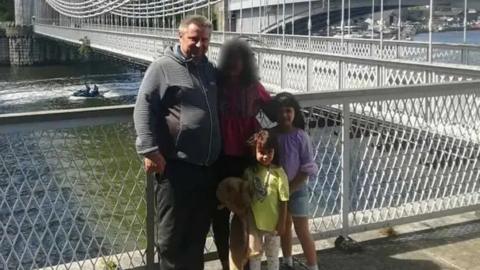 Bartlomiej Kuczynsk in a grey hoodie and black trousers standing on a riverside which has white railings. A moving boat is in the background travelling from right to left. His two young daughters, one wearing purple and one wearing yellow, are standing with him. A woman is pictured, whose face is blurred out.