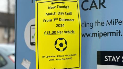 A yellow plastic sign details near parking charges. It has been placed on another larger blue parking sign. It reads: "New football match day tariff from 3rd December 2024, £15 per vehicle."