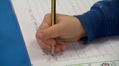 A child wearing a blue sweatshirt is writing in an exercise book with a yellow and black striped pencil.
