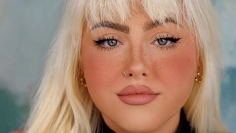 A close-up shot of Megan Lafferty, a young woman with blonde hair and freckled skin wearing glamorous make-up, a black jumper and gold earrings.